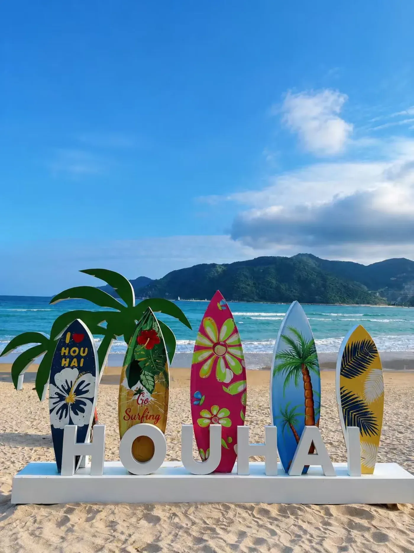 Wooden surfboard decorations spelling out HOUHAI on the beach, with the bay and hills behind